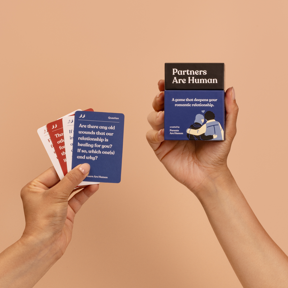 One hand holding a fan of Partners Are Human cards, the front card reading “Are there any old wounds that our relationship is healing for you? If so, which one(s) and why?”, and another hand holding the Partners Are Human box.