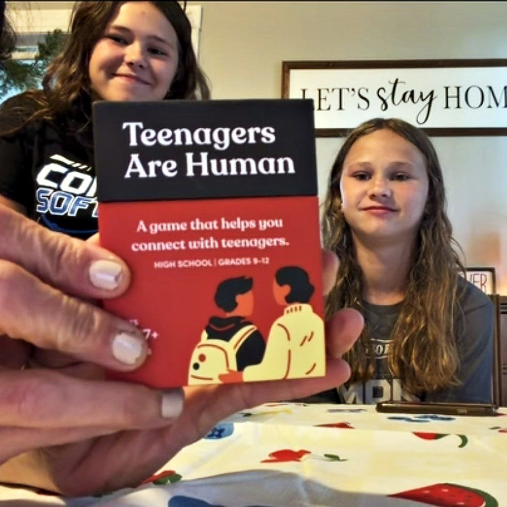 A parent holds a Teenagers Are Human box toward the camera while two teenage girls smile at the table