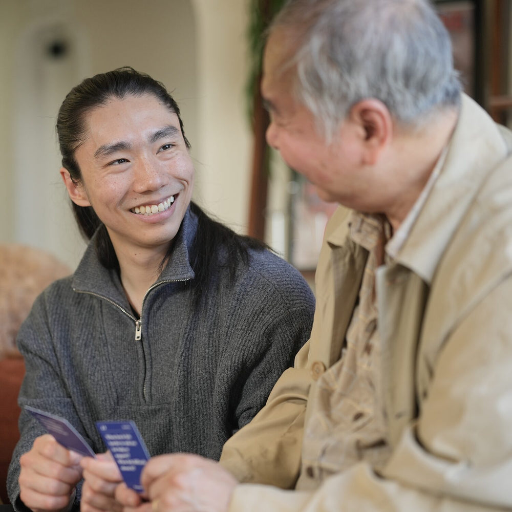 A son and his father laughing together on a sofa, each holding Parents Are Human question cards